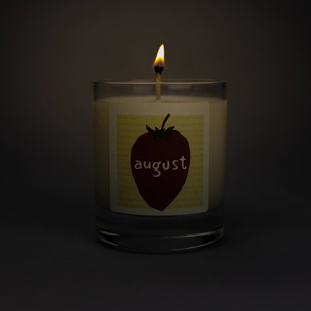 Product photo of lit scented soy candle August in the dark, made by BONKI.  Candle in glass jar. Yellow label with strawberry and writing "August".
