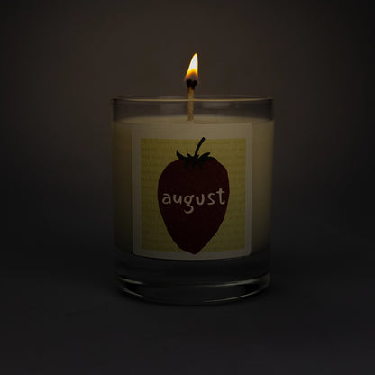 Product photo of lit scented soy candle August in the dark, made by BONKI.  Candle in glass jar. Yellow label with strawberry and writing "August".