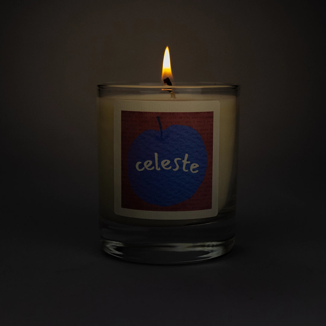 Product photo of lit scented soy candle Celeste in the dark, made by BONKI. Candle in glass jar. Pink label with purple peach and writing "Celeste".