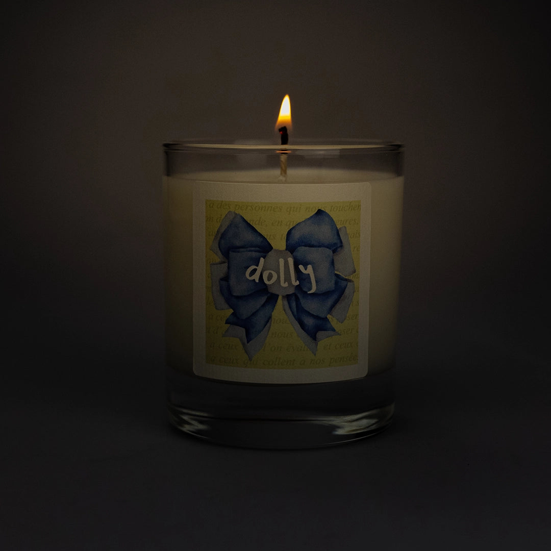 Product photo of lit scented soy candle Dolly in the dark, made by BONKI. Candle in glass jar. Yellow label with blue ribbon and writing "Dolly".