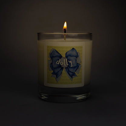 Product photo of lit scented soy candle Dolly in the dark, made by BONKI. Candle in glass jar. Yellow label with blue ribbon and writing "Dolly".
