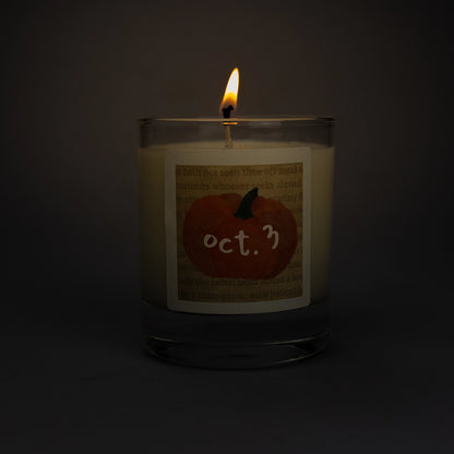 Product photo of lit scented soy candle Oct. 3 in the dark, made by BONKI. Candle in glass jar. Beige label with orange pumpkin and writing "Oct. 3".