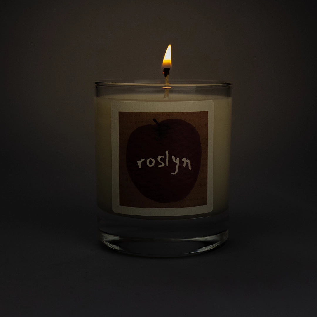 Product photo of lit scented soy candle Roslyn in the dark, made by BONKI. Candle in glass jar. Brown label with red apple and writing "Roslyn".
