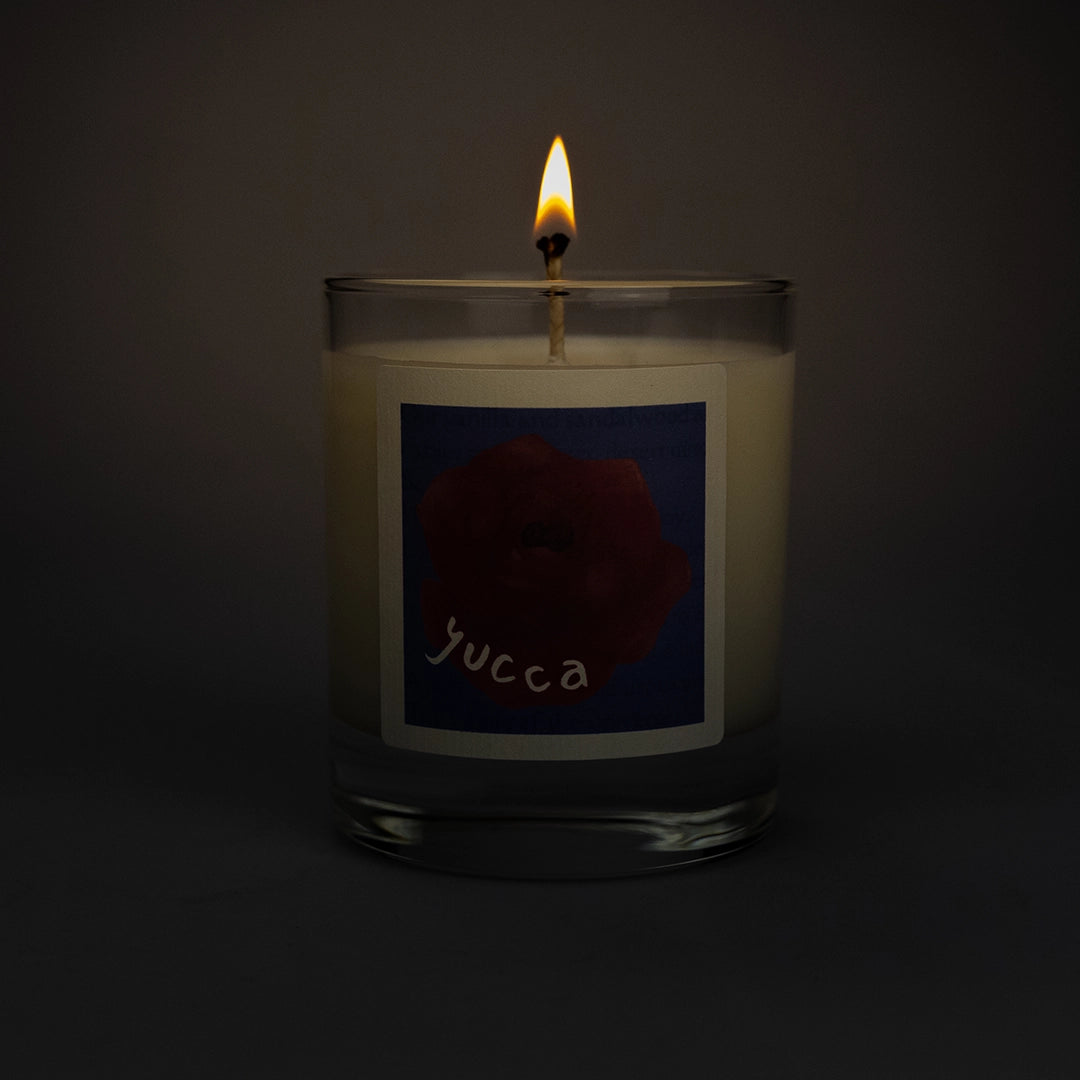Product photo of lit scented soy candle Yucca in the dark, made by BONKI. Candle in glass jar. Blue label with red rose and writing "Yucca".
