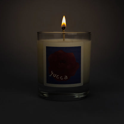Product photo of lit scented soy candle Yucca in the dark, made by BONKI. Candle in glass jar. Blue label with red rose and writing "Yucca".